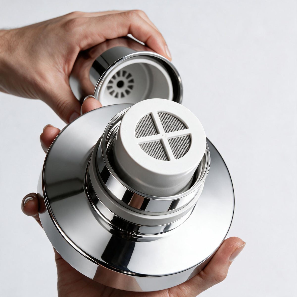 A close-up of a person's hands holding a polished chrome showerhead with a white mesh filter insert, shown against a clean light gray background.