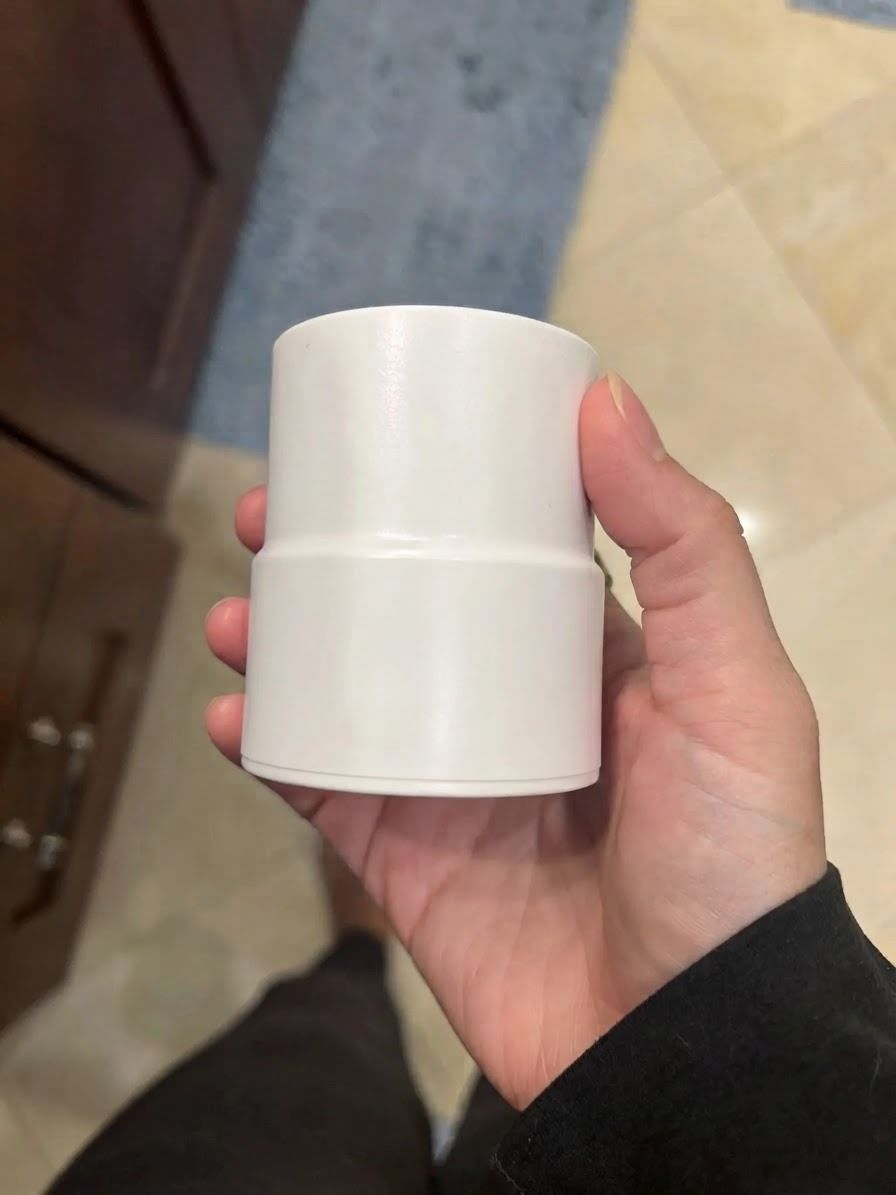 A hand holding a plain white cylindrical plastic pipe fitting against an indoor tiled floor and cabinet background.