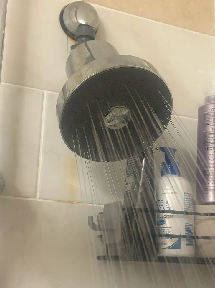 A chrome shower head with water running from it, mounted on a beige tiled wall. A small black shelf holding toiletries is visible below, with a purple bottle and white bottles nearby.